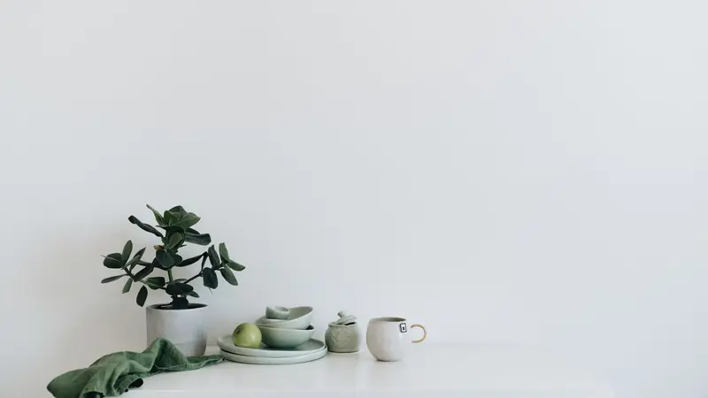 A calm still life of sage ceramics, a pear and a plant on a white surface.