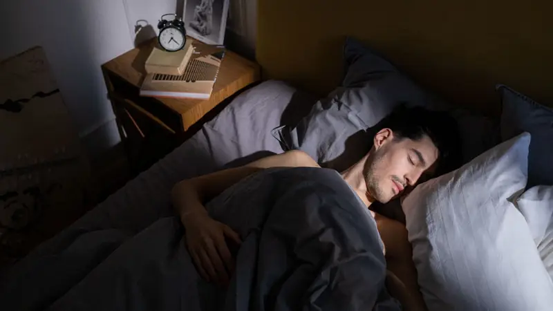 A person asleep in bed in a dark room, lit by a warm bedside lamp.